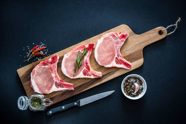 Raw pork loin chops shot from above on dark background Three raw pork loin chops shot from above on black background. The pork chops are on a wooden cutting board and some ingredients and spices like peppercorns, rosemary, marine salt, garlic and tomatoes are all around them. Predominant colors are red and brown. XXXL 42Mp studio photo taken with Sony A7rii and Sony FE 90mm f2.8 macro G OSS lens meat chop stock pictures, royalty-free photos & images