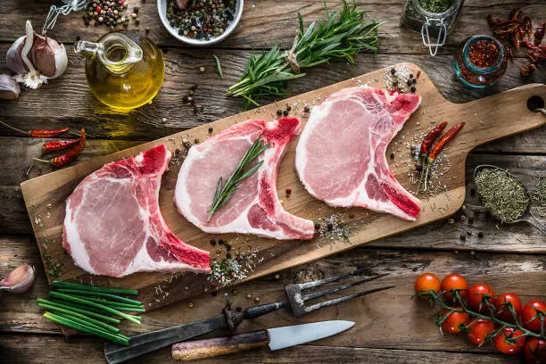 Three raw pork loin chops shot from above on rustic wooden table. The pork chops are on a wooden cutting board and some ingredients and spices like peppercorns, rosemary, marine salt, garlic and tomatoes are all around them. Predominant colors are red and brown. XXXL 42Mp studio photo taken with Sony A7rii and Sony FE 90mm f2.8 macro G OSS lens