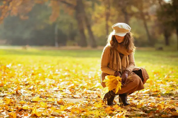 Hello autumn. trendy woman in sweater, skirt, hat, gloves and scarf outdoors in the autumn park gathering leaves.