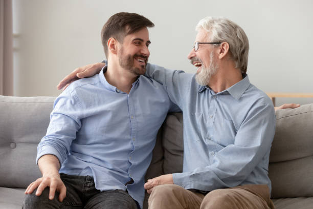 Happy old dad embracing young son talking laughing on sofa Happy old senior dad embracing young adult grown son talking laughing sit on sofa at home at reunion meeting, two generations male family relaxing having fun enjoy bonding together on fathers day father son grandfather fathers day stock pictures, royalty-free photos & images