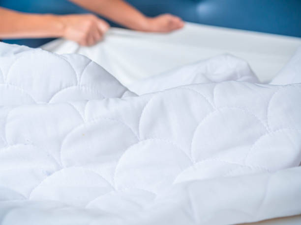 Selective focus at crumpled and wrinkle white bed topper on the bed in the bedroom with blurry woman 's hand pull the bed cover. Housework preparation before laundry process. stock photo
