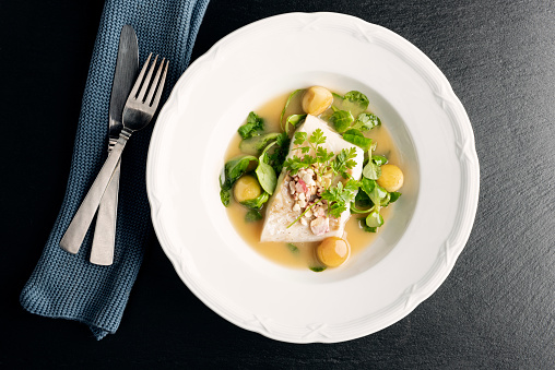 Overhead dish of cod fillet in a beurre blanc sauce with apple balls, and sugar salted apple chips, parsley and watercress at the pass before serving. Colour, horizontal  with some copy space, photographed on location at a restaurant on the island of Moen in Denmark.