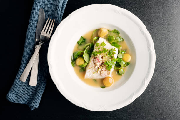 Cod fillet with beurre blanc sauce and apple. Overhead dish of cod fillet in a beurre blanc sauce with apple balls, and sugar salted apple chips, parsley and watercress at the pass before serving. Colour, horizontal  with some copy space, photographed on location at a restaurant on the island of Moen in Denmark. serving dish stock pictures, royalty-free photos & images