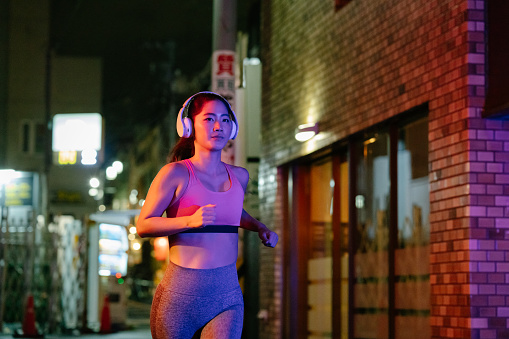 A young female athlete is running in the street while listening to music with bluetooth wireless headphones, The streets are wet after the rain. She is lit by vibrant neon colored lights.