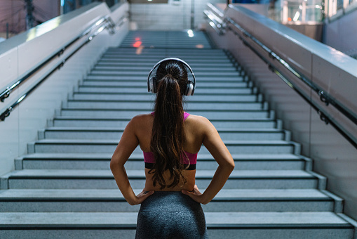 A young female sports person is standing confidently in font of steps while listening to music with a wireless bluetooth headphones.
