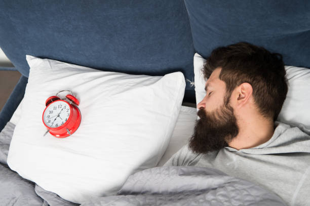 istock Man bearded hipster sleepy face in bed with alarm clock. Problem with early morning awakening. Get up with alarm clock. Overslept again. Tips for waking up early. Tips for becoming an early riser 1186355538