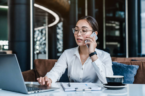 istock Smiling asian female talking on mobile phone while sitting on desk with laptop 1186357070