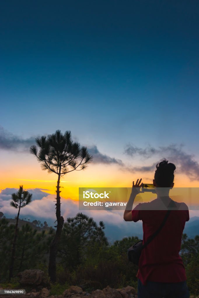 Woman shoots cloudy sunset view in the mountains through a smartphone. Image of an Asian/Indian young woman in an outdoor scene takes photos of the mountains in cloudy sunset through a smartphone in Solan, Himachal Pradesh, India. Himachal Pradesh Stock Photo