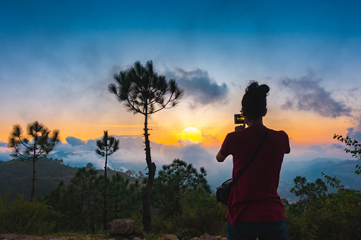Image of an Asian/Indian young woman in an outdoor scene takes photos of the mountains in cloudy sunset through a smartphone in Solan, Himachal Pradesh, India.