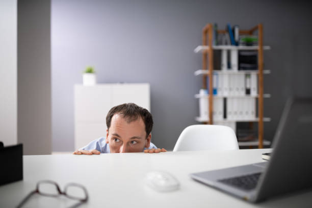 afraid businessman peeking from the edge of desk in office - hiding fear men peeking fotografías e imágenes de stock