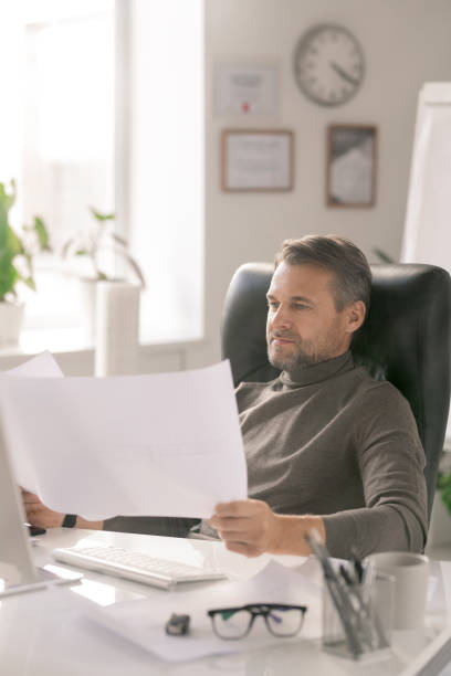 istock Mature confident architect looking at sketches on papers while sitting by desk 1186381425