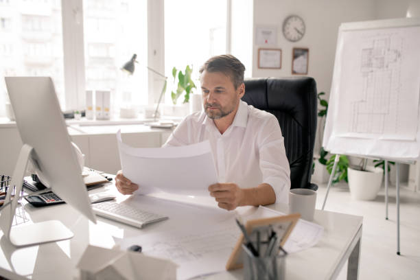istock Mature serious architect in white shirt checking sketches by workplace 1186381433
