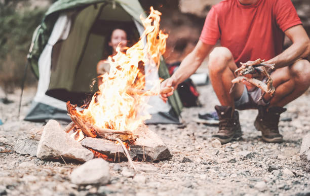 Happy couple making fire while camping in wild forest - Young people having fun traveling and camping next bonfire - Travel vacation lifestyle concept