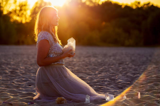 A stunning woman in a white off-shoulder dress sits on a picnic blanket illuminated by glowing fairy lights, enjoying a peaceful evening by the ocean at sunset. Perfect for lifestyle and romance themes.