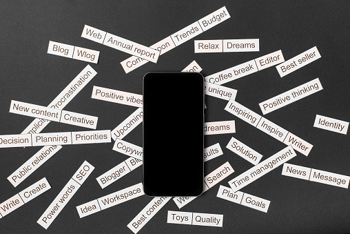 Smartphone surrounded by a cloud of words on business themes cut out of paper on a gray background,