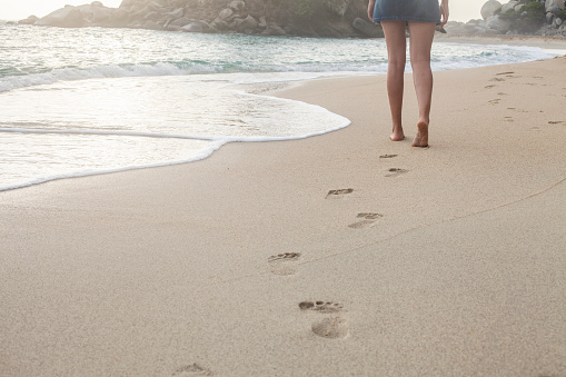 Latin woman with short jean skirt walks the sand on the beach leaving footprints on her way