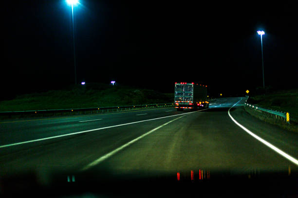 Car point of view looking through windshield at a solitary late night expressway speeding semi tractor trailer truck hauling container freight on an otherwise deserted midnight highway. The car is entering the multiple lane highway to merge from the right after a curving entrance ramp road.