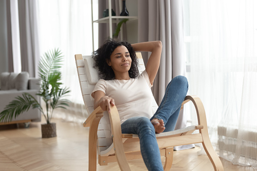 African woman in casual clothes leaned on wooden rocking chair resting in living room, female relax enjoy calmness breathing fresh air, spend free time at home, no stress, modern furniture ad concept
