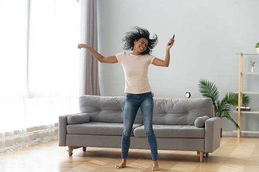 Full length african woman shake head raise hands holds remote control stereo music system or cell phone enjoy playlist favourite song dance in living room, bliss mood hobby weekend activities concept