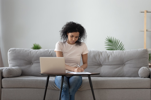 African woman sit on sofa at coffee table holding pen writing working from home, noting writing down, housewife planning week write to-do list, student learning preparing university admission concept