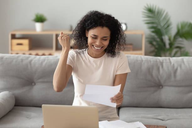 Woman holding paper reading letter feels happy celebrate loan approval African woman sitting on couch at home holding paper reading letter from bank feels happy about loan approval, overjoyed american female makes yes gesture with hand celebrate getting new job concept college acceptance letter stock pictures, royalty-free photos & images