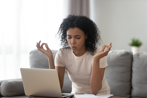 African woman sit on couch near laptop take break reduce stress do yoga meditation exercise to calm down self control get rid of negative emotions, bad e-mail, difficult task, problems at work concept