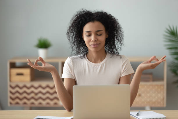 African woman meditating sitting at desk in front of laptop African woman meditating sit at desk in front of pc, serene mixed-race female closed eyes folded fingers mudra symbol do exercise practising yoga reducing anxiety stress positive frame of mind concept break time stock pictures, royalty-free photos & images