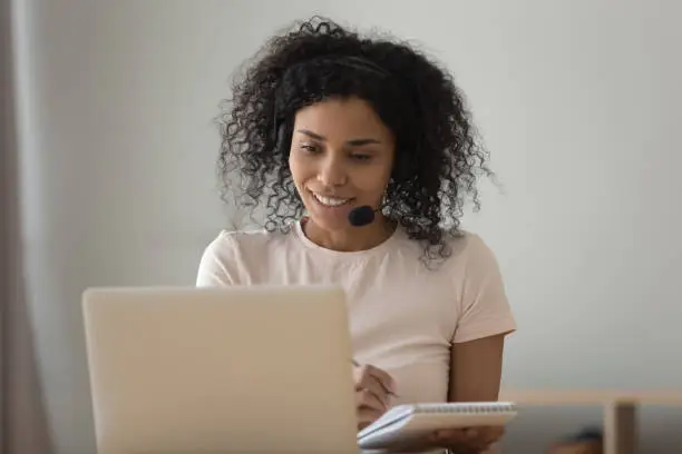 African woman interpreter in headset working from home online translation job, girl improve foreign language skills look at pc learns repeat words memorizes them, do task use internet website concept