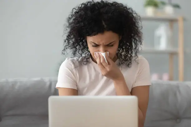 Photo of African woman holding handkerchief blowing runny nose working on laptop