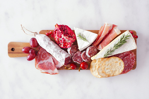 Serving board of assorted meats, cheeses and appetizers. Top view on a white marble background.