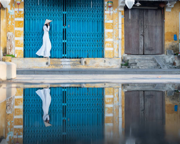 Young Vietnamese Woman in White Tunic  Walking in Front of a Turquoise Door Young Vietnamese woman wearing a white ao dai or traditional garment with a non la or straw hat walking in front of a large turquoise door with grate. ao dai stock pictures, royalty-free photos & images