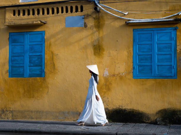 Vietnamese Woman in Traditional White Garment Walking in Front of a Gold Stucco Building Vietnamese woman wearing a white ao dai or traditional tunic and pant suit and a non la or conical straw hat while walking in front of a gold stucco building with brilliant turquoise shuttered windows. ao dai stock pictures, royalty-free photos & images