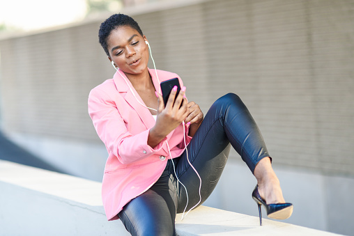 Black businesswoman sitting outdoors speaking via videoconference with her smart phone. African american female wearing suit with pink jacket.