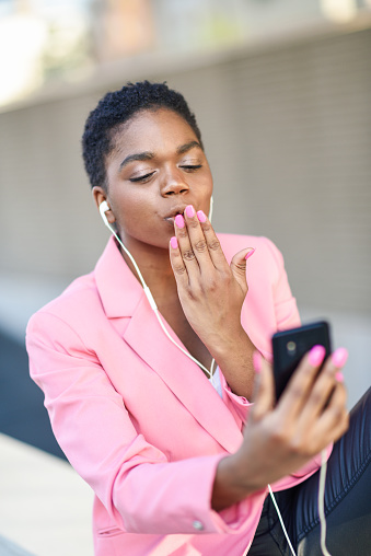 Black businesswoman sitting outdoors speaking via videoconference with her smartphone. African american female wearing suit with pink jacket.