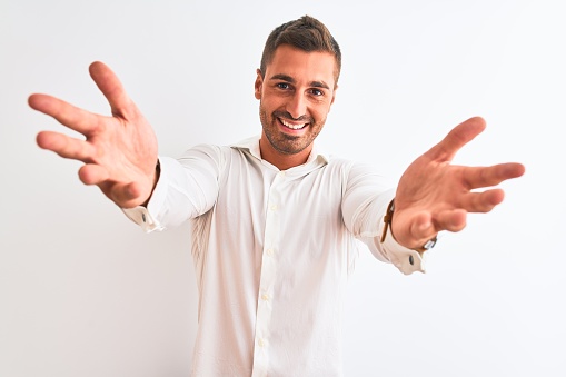 Young handsome business man wearing elegant shirt over isolated background looking at the camera smiling with open arms for hug. Cheerful expression embracing happiness.