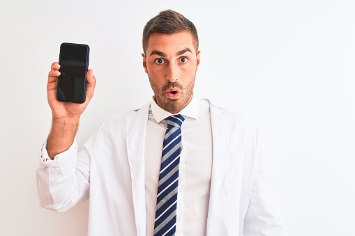 Young scientist man showing smartphone screen over isolated background scared in shock with a surprise face, afraid and excited with fear expression