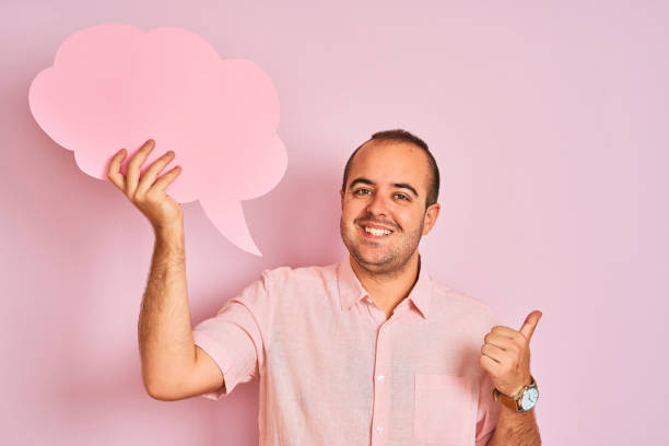 istock Young man holding cloud speech bubble standing over isolated pink background pointing and showing with thumb up to the side with happy face smiling 1186468164