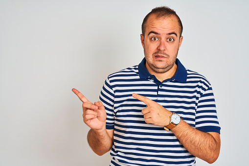 Young man wearing casual striped polo standing over isolated white background Pointing aside worried and nervous with both hands, concerned and surprised expression