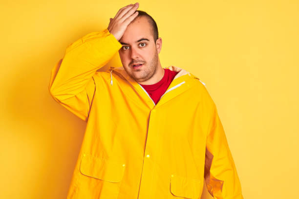istock Young man wearing rain coat standing over isolated yellow background surprised with hand on head for mistake, remember error. Forgot, bad memory concept. 1186469367