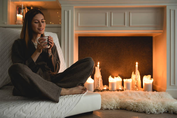 belle femme s'asseyant confortable à côté de la cheminée avec des bougies, retenant une tasse avec l'ornement de noel. - carpet caucasian domestic room home interior photos et images de collection