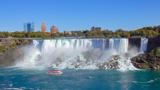 View of tour boat cruising at the base of misty Niagara Falls as seen from the Skylon Tower. Whitewater waterfalls American & Bridal Veil Falls, touristic hotels and New York skyline in the background