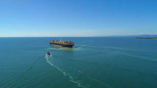 AERIAL: Big international freight ship setting out to sea after stopping at large international ocean port. Freight ship carrying containers is accompanied by boats drifting towards industrial harbor.