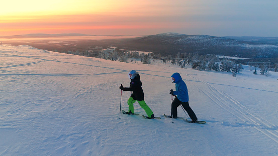 AERIAL: Active couple in warm winter ware snowshoeing on snowy mountain at sunrise. Man tour guide and woman snowshoe hiking at winter sunset. Travelers snowshoe exploring snowy Lapland mountains