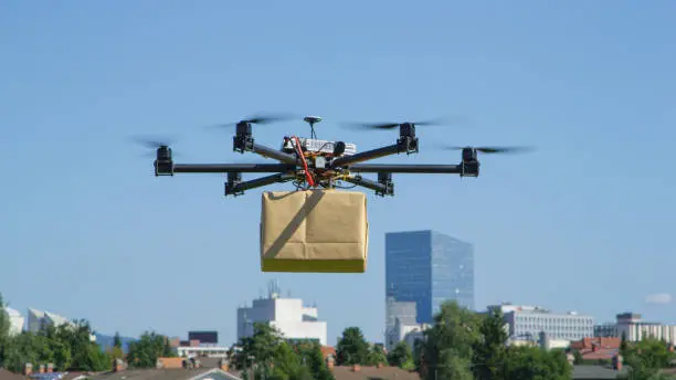 CLOSE UP: UAV drone delivery. Multicopter flying big brown package into city. Drone delivering post package to your home. Futuristic shipment by helicopter drone. Multirotor logistics and transport.