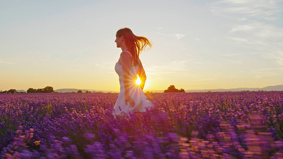Cheerful young woman in white dress running through beautiful purple lavender field at golden sunset in summer