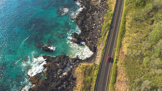 AERIAL: Red convertible driving along the beautiful coastal road above the rocky ocean cliffs and crystal clear sea water splashing into the rocks
