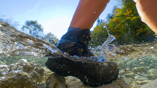 HALF-HALF, LENS FLARE: Female trekker crossing river rushing down rocky riverbed. Picturesque hike down mountain valley in soothing autumn sunlight. Cold river stream flowing down pebbles and rocks.