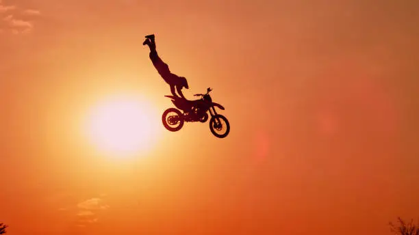 SILHOUETTE: Pro motocross rider riding fmx motorbike, jumping big air kicker performing extreme stunt. Professional biker jumps no hander superman trick over orange sunset sky