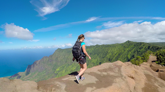 Sportswoman walking on the edge of high mountain ridge overlooking vast jungle valley and blue ocean bay. Young woman on summer vacation hiking in Hawaiian mountains on a dirt path with stunning view
