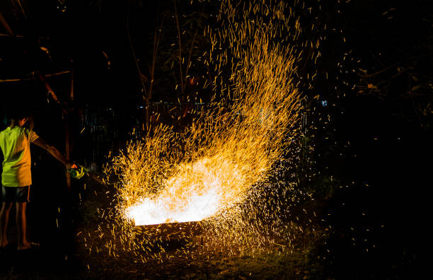 A kid stands by a furnace using a portable blower fanning the flames creating sparks in the process